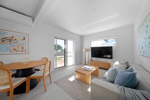 A modern living room with a dining area, a flat-screen TV, and a large window. - Esplanade Apartments | Ocean View Beachfront | West Beach (West Beach)
