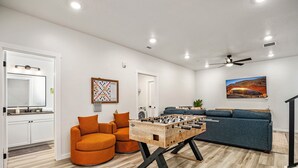 A modern living room with a foosball table, a sofa, a dining table, and a kitchen area with a sink and mirror. - Desert Haven, Private Spa, Community Amenities w/ Water Slide (Hurricane)