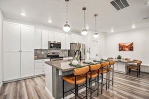 A modern kitchen with white cabinets, stainless steel appliances, a central island with bar stools, and a dining area with a table and chairs. - Desert Haven, Private Spa, Community Amenities w/ Water Slide (Hurricane)