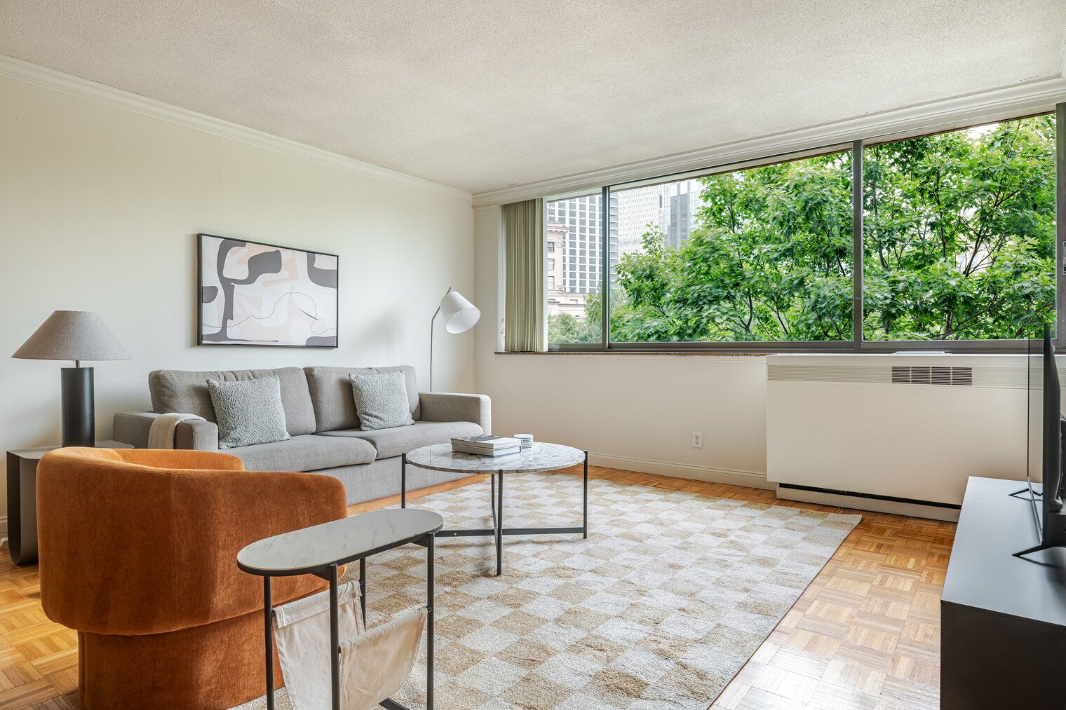 A modern living room with a sofa, coffee table, and a view of trees through large windows.
