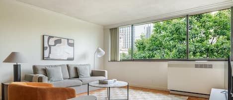 A modern living room with a sofa, coffee table, and a view of trees through large windows.