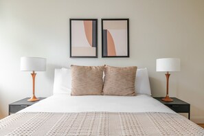 A hotel room with a bed, two nightstands, two lamps, two pillows, and two abstract paintings on the wall. - Blueground | Fenway–Kenmore, fitness, nr Fenway Park (Boston)