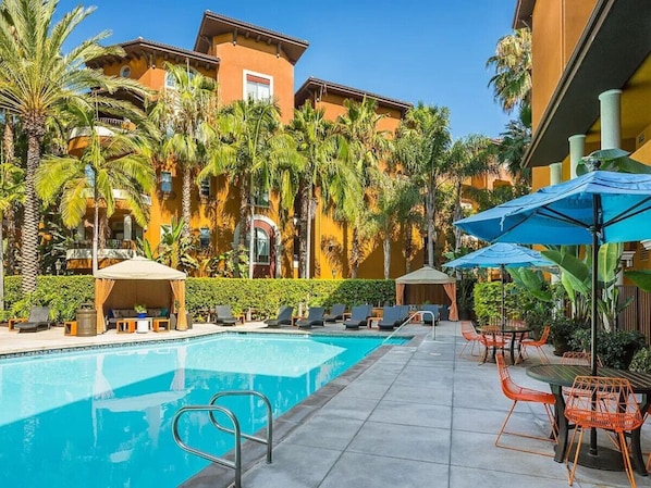 A swimming pool with lounge chairs and umbrellas, surrounded by palm trees and a building with balconies.