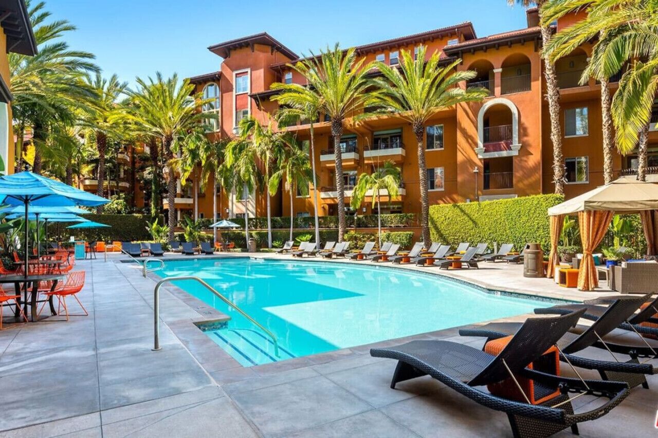 Une piscine entourée de palmiers, avec des chaises longues et des parasols, et un immeuble à plusieurs étages doté de balcons.