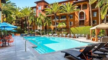 Une piscine entourée de palmiers, avec des chaises longues et des parasols, et un immeuble à plusieurs étages doté de balcons.