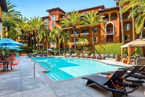 Una piscina con sillas de descanso y sombrillas, rodeada de palmeras y un edificio de varios pisos con balcones.