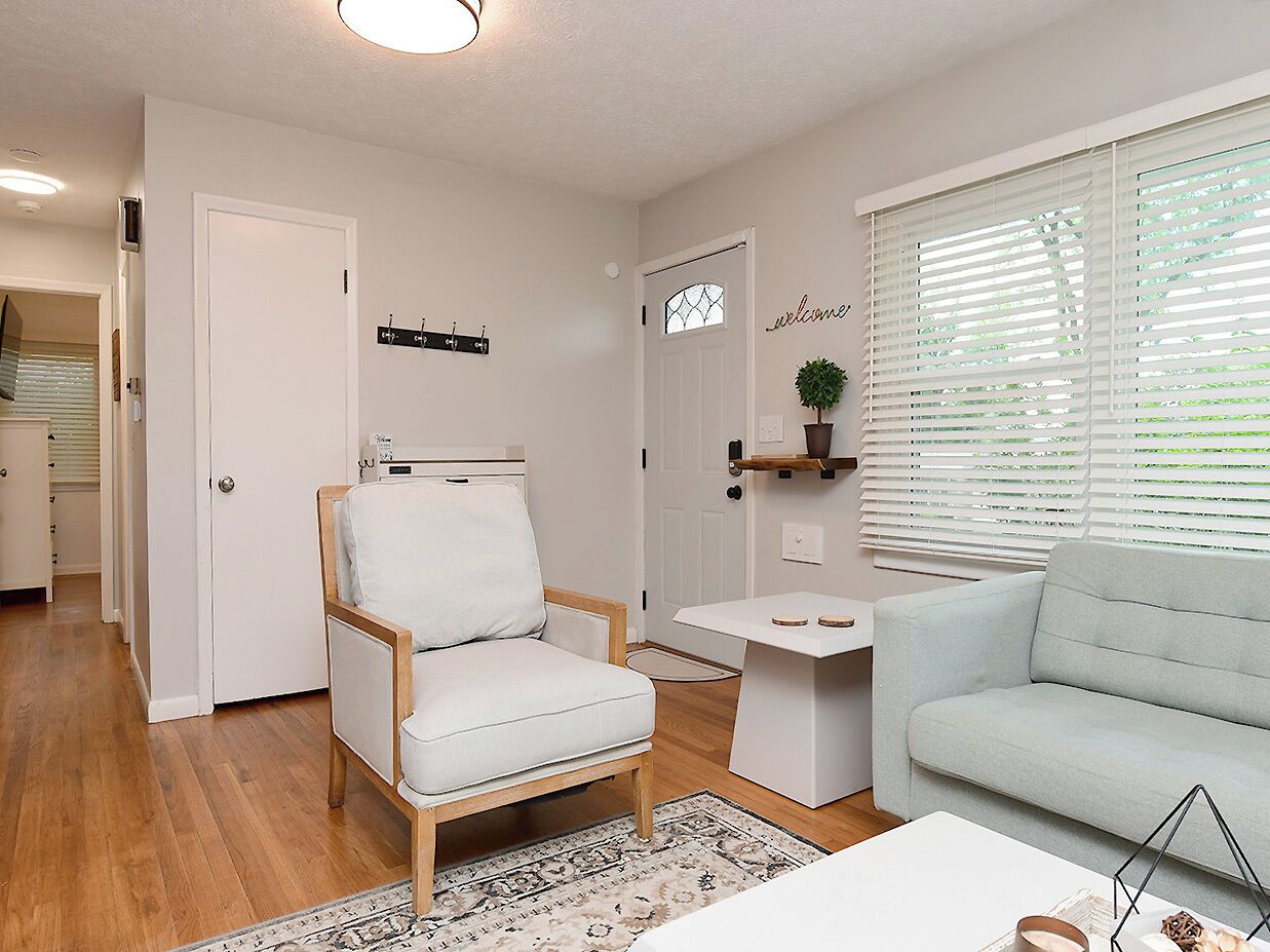 A living room with a white armchair, a light blue sofa, a small white table, and a window with blinds.