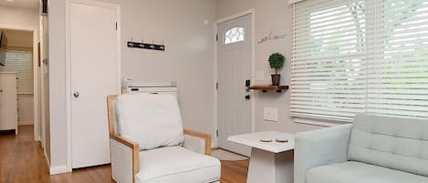 Una sala de estar con un sillón blanco, un sofá azul claro, una mesita pequeña y un ventanal con persianas.