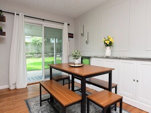 A dining area with a wooden table and benches, a sliding glass door leading to a patio, and white cabinetry. - Cozy Aksarben Gem 2BR • Quiet • Fenced Yard (Omaha)