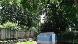 Pieni, kaltevakattoinen varasto sijaitsee nurmikon peitossa olevalla pihalla, ja sen ympärillä on puinen aita sekä puita.