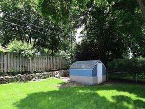 A small shed with a sloped roof in a grassy yard, surrounded by a wooden fence and trees. - Cozy Aksarben Gem 2BR • Quiet • Fenced Yard (Omaha)