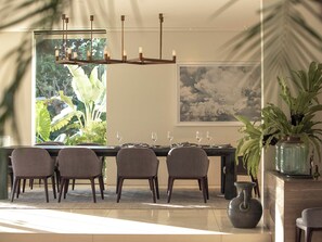 A dining area with a table set for four, chairs, a chandelier, and a large window with a view of greenery.