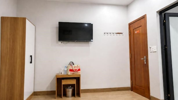 A room with a television mounted on the wall, a wooden wardrobe, a small table with a bag, and a door. - Hung Phat Hotel Nghe An  (Hung Nguyen)