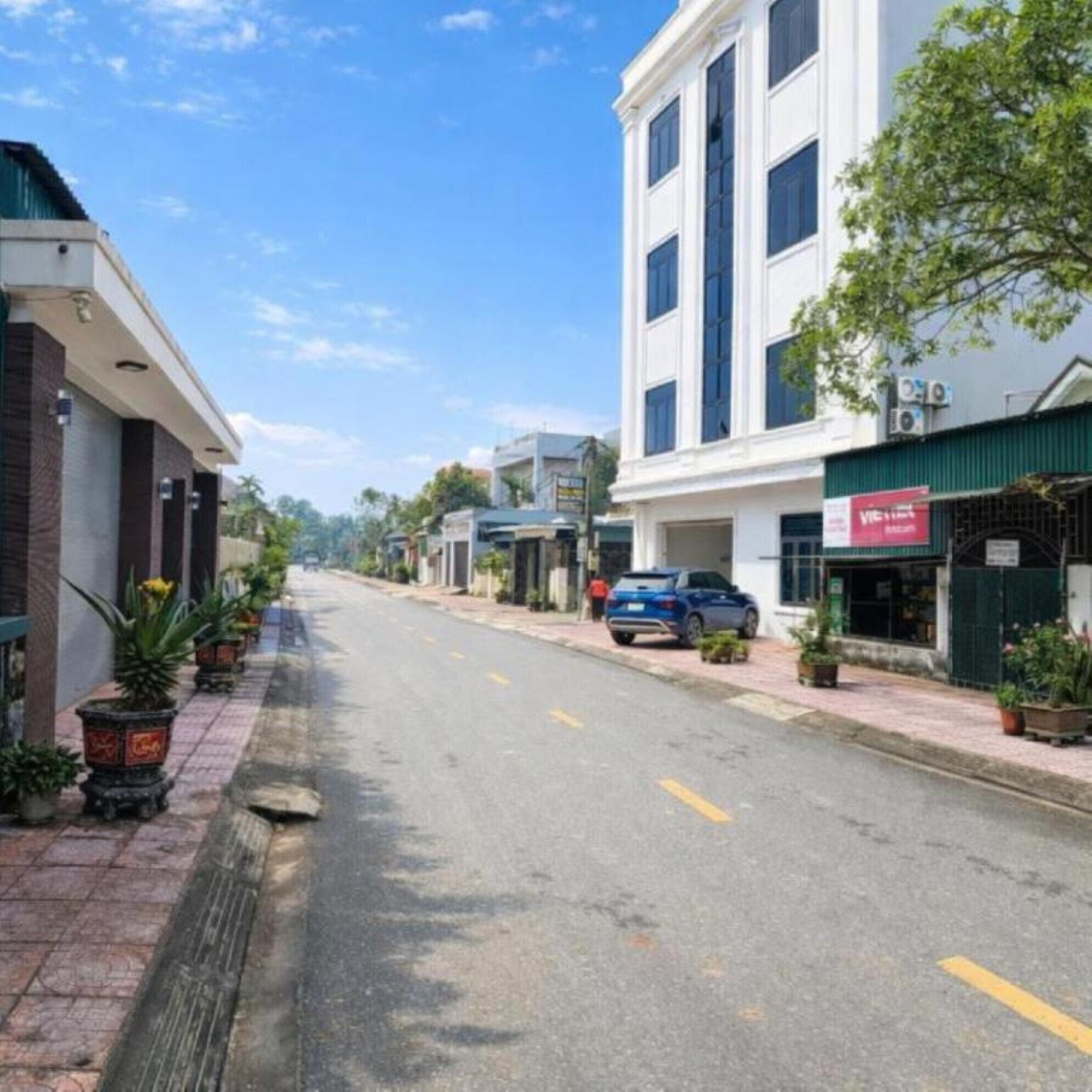 A street with buildings, a parked car, and potted plants along the sidewalk.