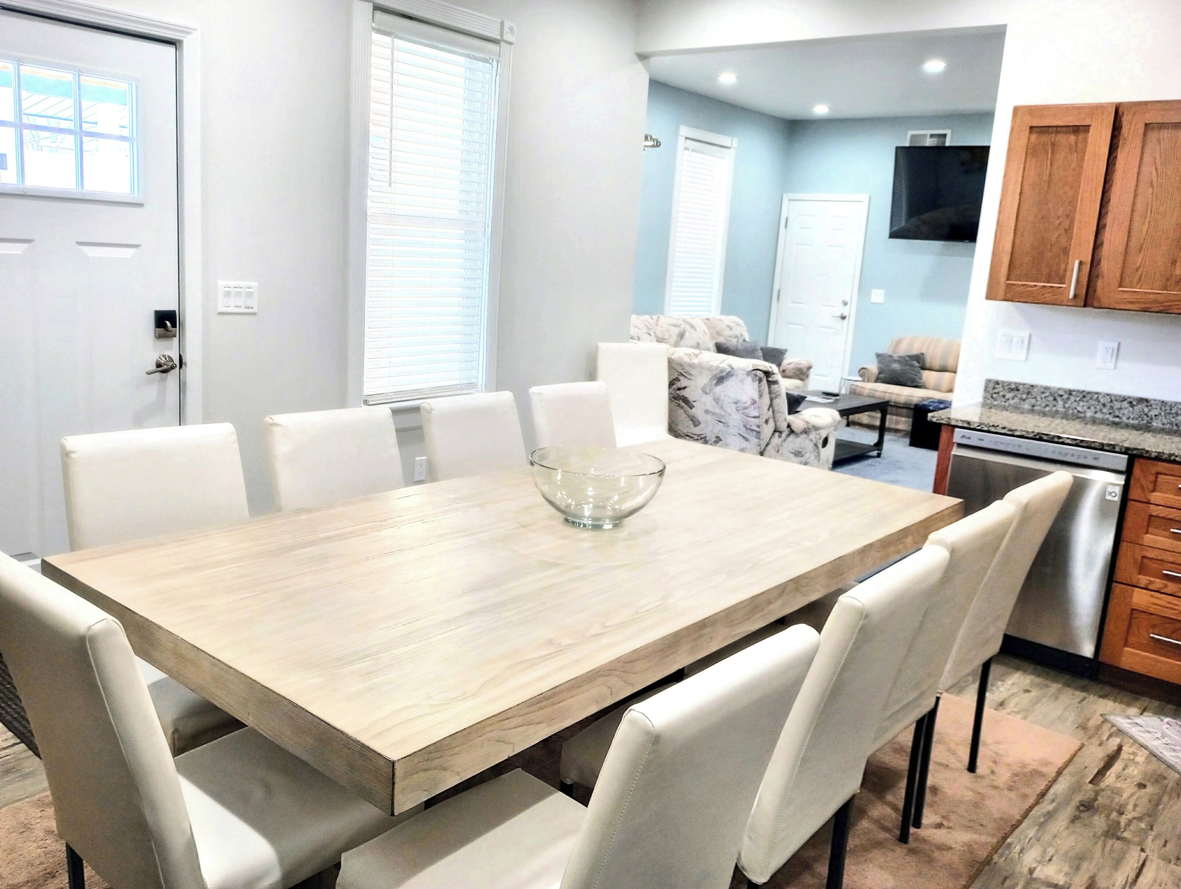 A dining area with a wooden table and white chairs, a kitchen with granite countertops, and a living area with a sofa and TV.
