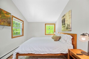 A bedroom with a large bed, two windows, a nightstand, a lamp, and a painting on the wall. - TreeTop Chalet 4BR & Pool - Upper Mtn Road Stowe (Stowe)