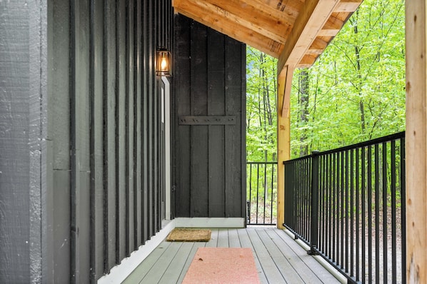 A wooden deck with a black metal railing, a door with a light fixture, and a view of a green forest. - TreeTop Chalet 4BR & Pool - Upper Mtn Road Stowe (Stowe)