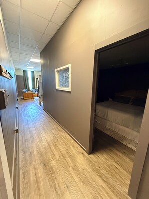 Ein Hotelkorridor mit einem Schlafraum mit Etagenbetten rechts, Holzboden und einem Fenster mit einem gemusterten Rollo.