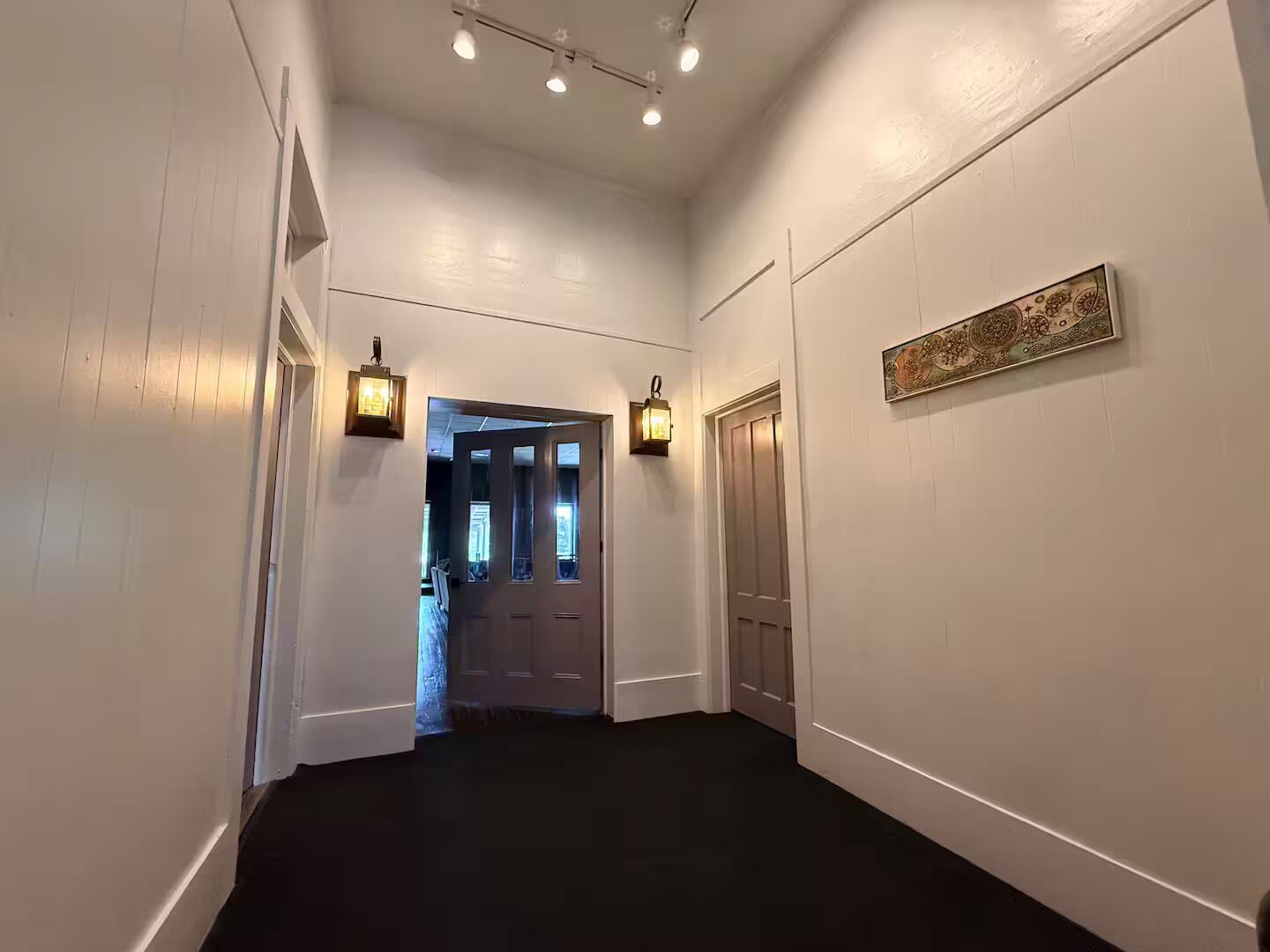Un couloir aux murs blancs, au sol noir et doté de deux portes. Des luminaires sont fixés aux murs de part et d’autre du couloir.