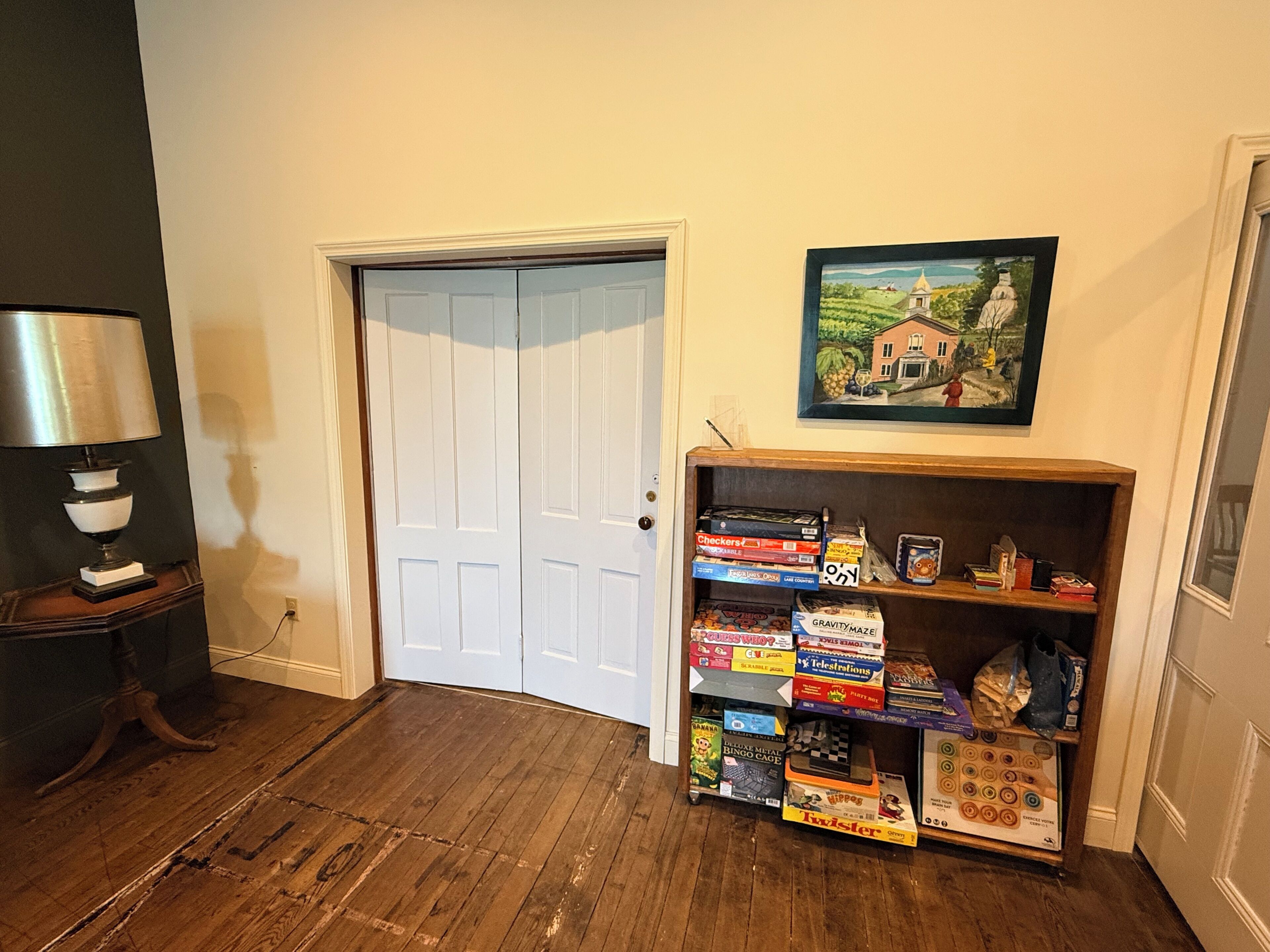 A room with a wooden floor, a white door, a lamp on a table, and a bookshelf with various board games.