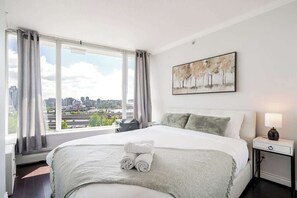 Ein Hotelzimmer mit einem großen Bett, Blick auf die Stadt, einem Gemälde an der Wand und einem Nachttisch mit einer Lampe.