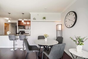 Eine moderne Küche mit Mikrowelle, Backofen und Pendelleuchten. Ein Essbereich mit rundem Tisch und vier Stühlen. Eine Uhr ist an der Wand.