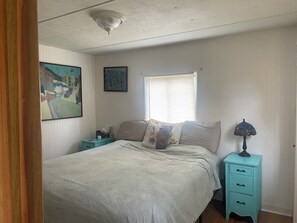 A bedroom with a bed, a nightstand, a lamp, a window with blinds, and two wall-mounted paintings.