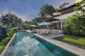 A swimming pool with lounge chairs and umbrellas, surrounded by lush greenery and a building with a covered patio. - Villa Bayu Uluwatu (denpasar)