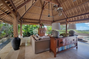 Un espace extérieur abrité, doté d’un toit de chaume, d’un canapé, d’une bibliothèque en bois et offrant une vue sur la verdure.