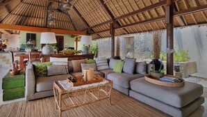 A spacious living area with a high thatched ceiling, a sofa set, a dining table, and a kitchen in the background. - Villa Bayu Uluwatu (denpasar)
