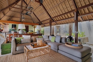 A spacious living area with a high thatched ceiling, a sofa set, a dining table, and a kitchen in the background. - Villa Bayu Uluwatu (denpasar)