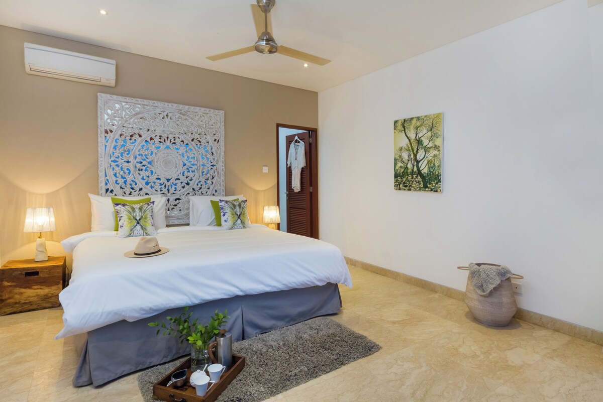 A bedroom with a bed, bedside lamps, a ceiling fan, a decorative wall piece, a door with a coat hanger, and a painting on the wall.