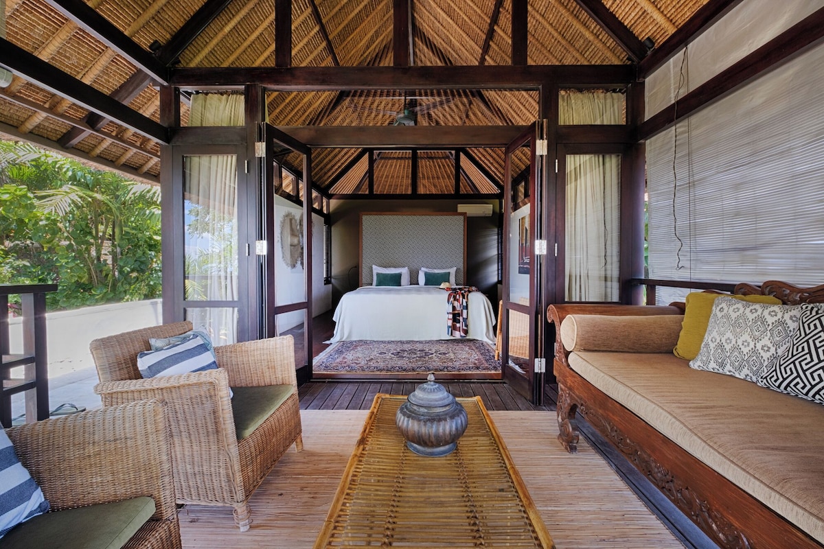 A room with a bed, a sofa, and two chairs. There is a window with curtains, a ceiling with a thatched roof, and a wooden floor.