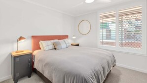 A bedroom with a bed, a nightstand, a desk, a ceiling fan, and a window with blinds. - Saltbush Beach Pad - Luxe apartment steps to the beach and cafes (Coffs Harbour)