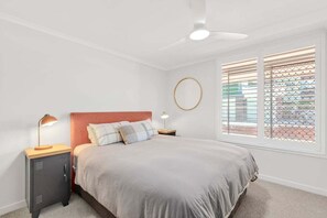 A bedroom with a bed, a nightstand, a desk, a ceiling fan, and a window with blinds. - Saltbush Beach Pad - Luxe apartment steps to the beach and cafes (Coffs Harbour)
