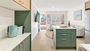 A modern kitchen with a central island, a dining area with a sofa, and a view of the outside. - Saltbush Beach Pad - Luxe apartment steps to the beach and cafes (Coffs Harbour)