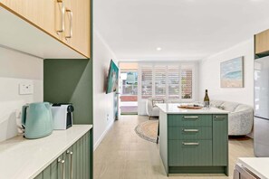 A modern kitchen with a central island, a dining area with a sofa, and a view of the outside. - Saltbush Beach Pad - Luxe apartment steps to the beach and cafes (Coffs Harbour)