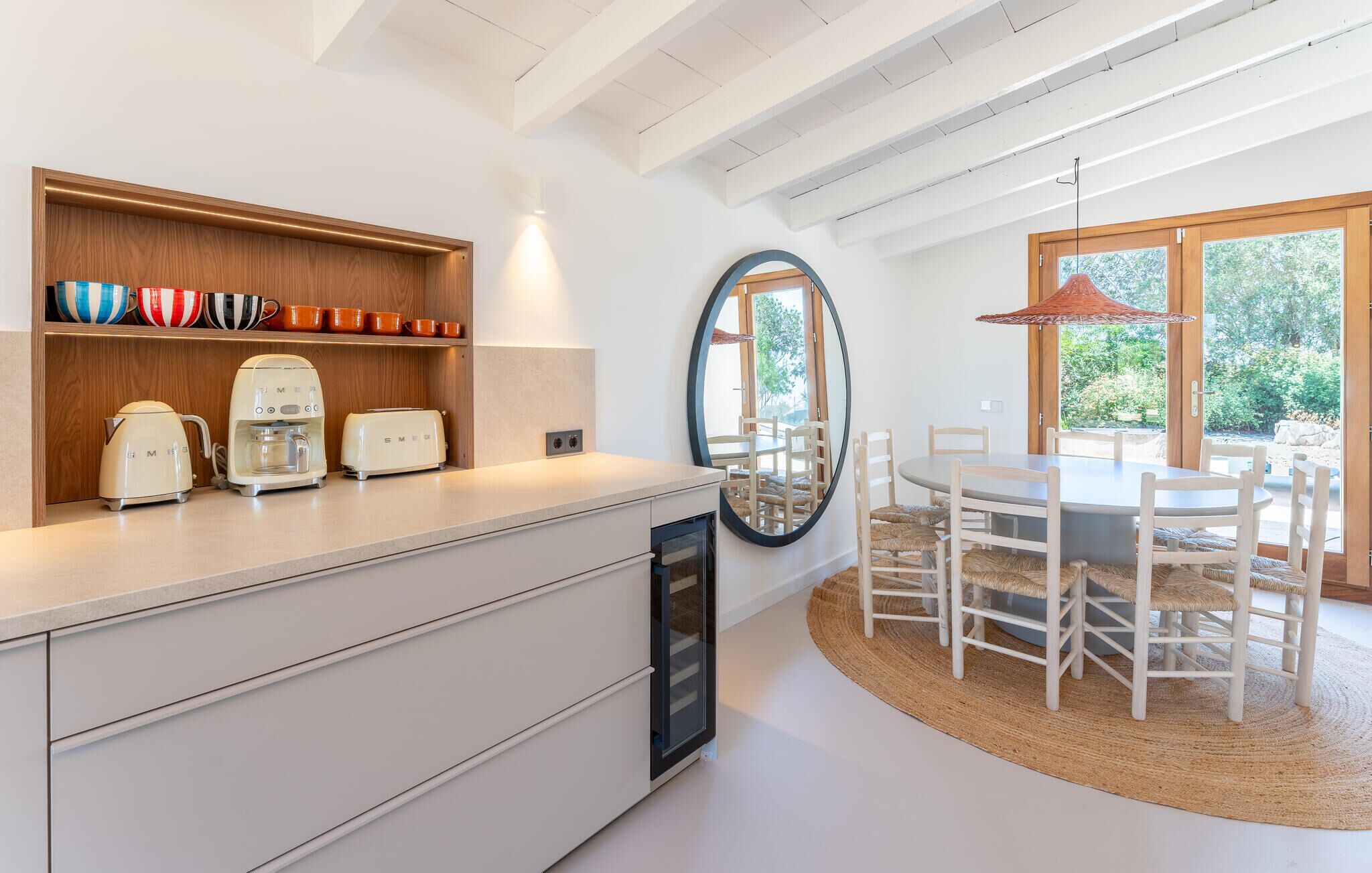 A modern kitchen with white cabinetry, a built-in coffee maker, and a toaster. There is a dining area with a round table and chairs, and a large mirror on the wall.