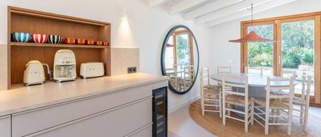 A modern kitchen with white cabinetry, a built-in coffee maker, and a toaster. There is a dining area with a round table and chairs, and a large mirror on the wall.