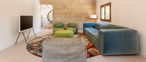Un salon moderne avec un canapé d’angle, des poufs verts et gris, un téléviseur à écran plat posé sur un meuble et un mur en pierre.