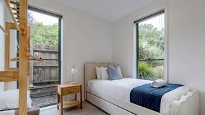 Ein Schlafzimmer mit Bett, Nachttisch, Lampe und Blick auf Grün hinter großen Fenstern.