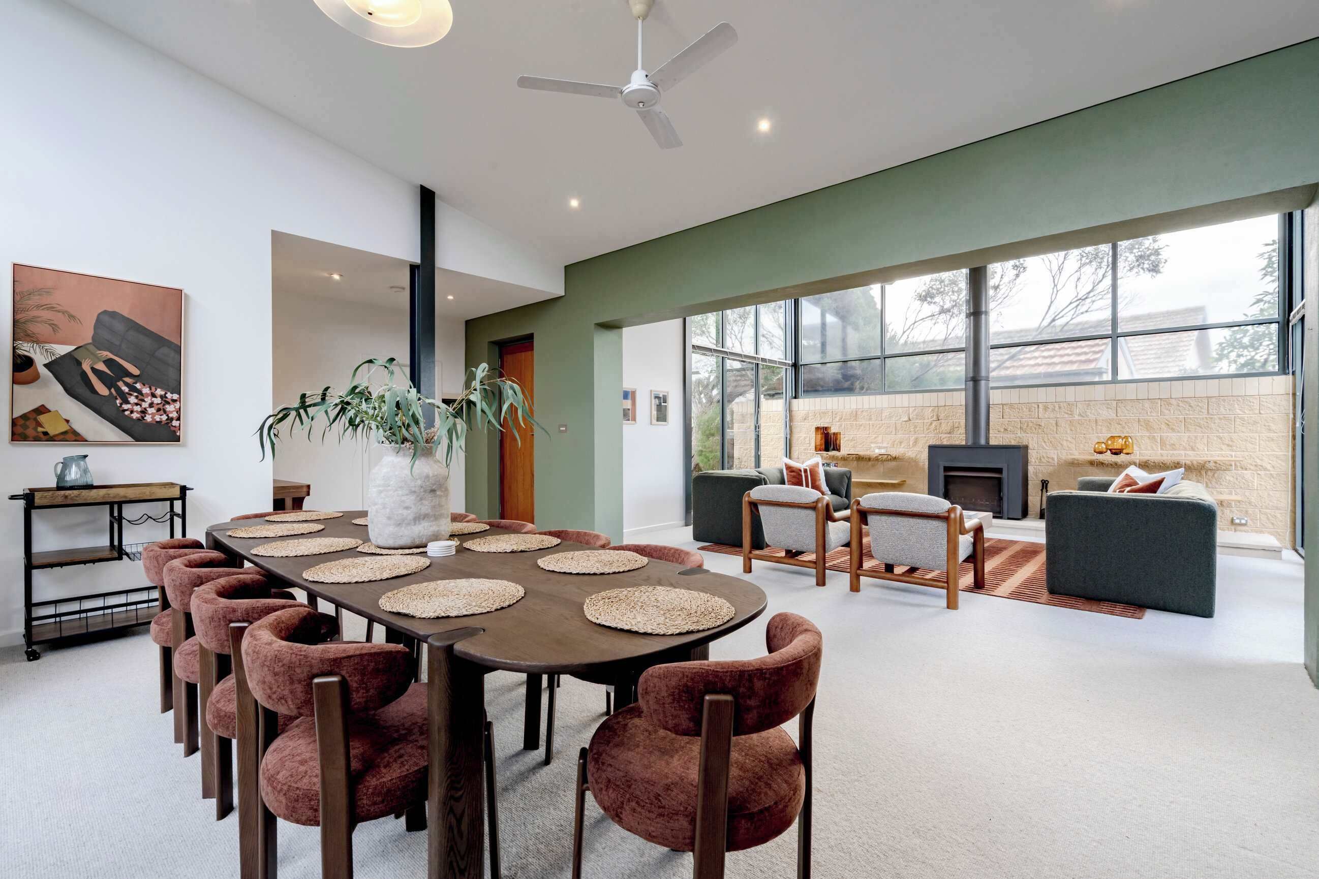 A modern living room with a dining area, large windows, a fireplace, and a ceiling fan.