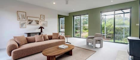 A modern living room with a sofa, coffee table, and armchairs, large windows, and artwork on the walls.