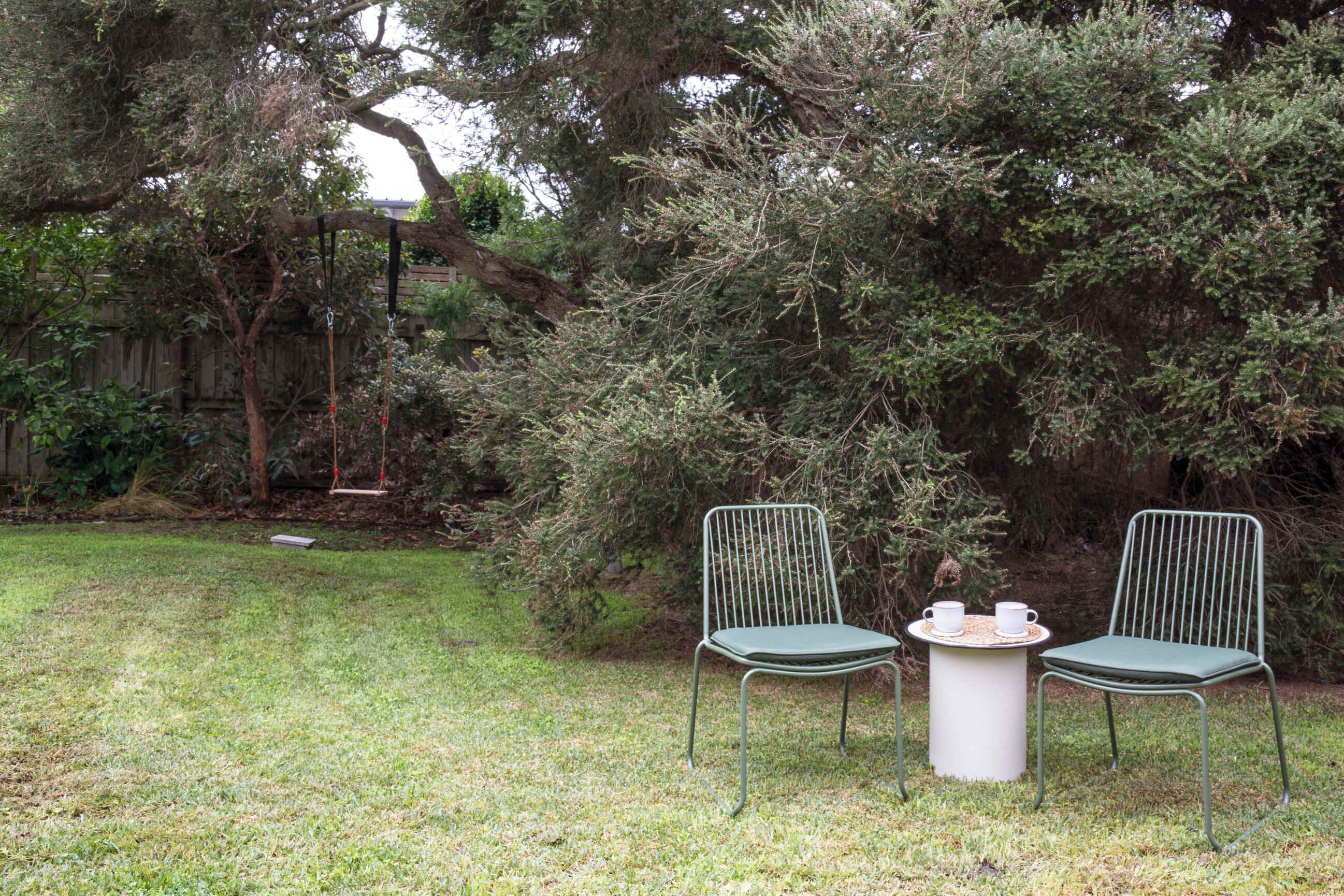 A grassy outdoor area with two metal chairs and a small table, surrounded by dense foliage and a wooden fence.