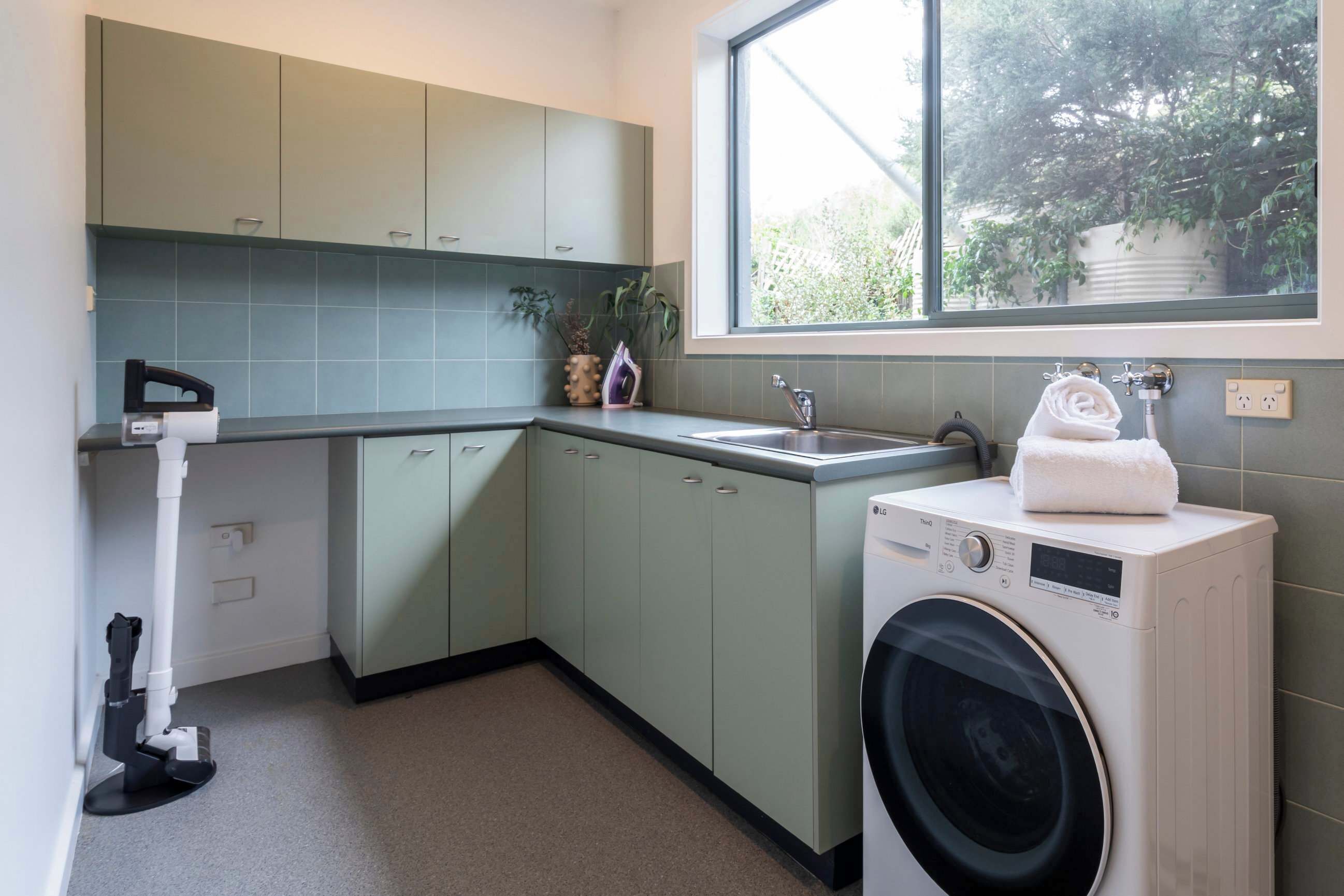 A compact kitchen with a washing machine, a sink, and a window with a view of greenery.