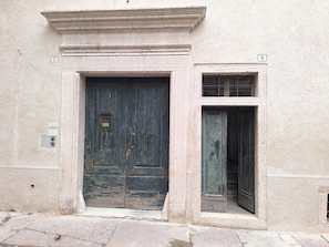 Fachada de un edificio desgastado por el tiempo, con una puerta principal grande de madera oscura y otra puerta más pequeña a la derecha.