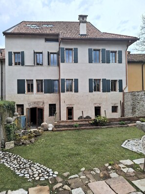 A two-story building with a tiled roof, multiple windows with shutters, and a small front garden with stepping stones and plants. - 101 - Fireplace Room (Belluno)