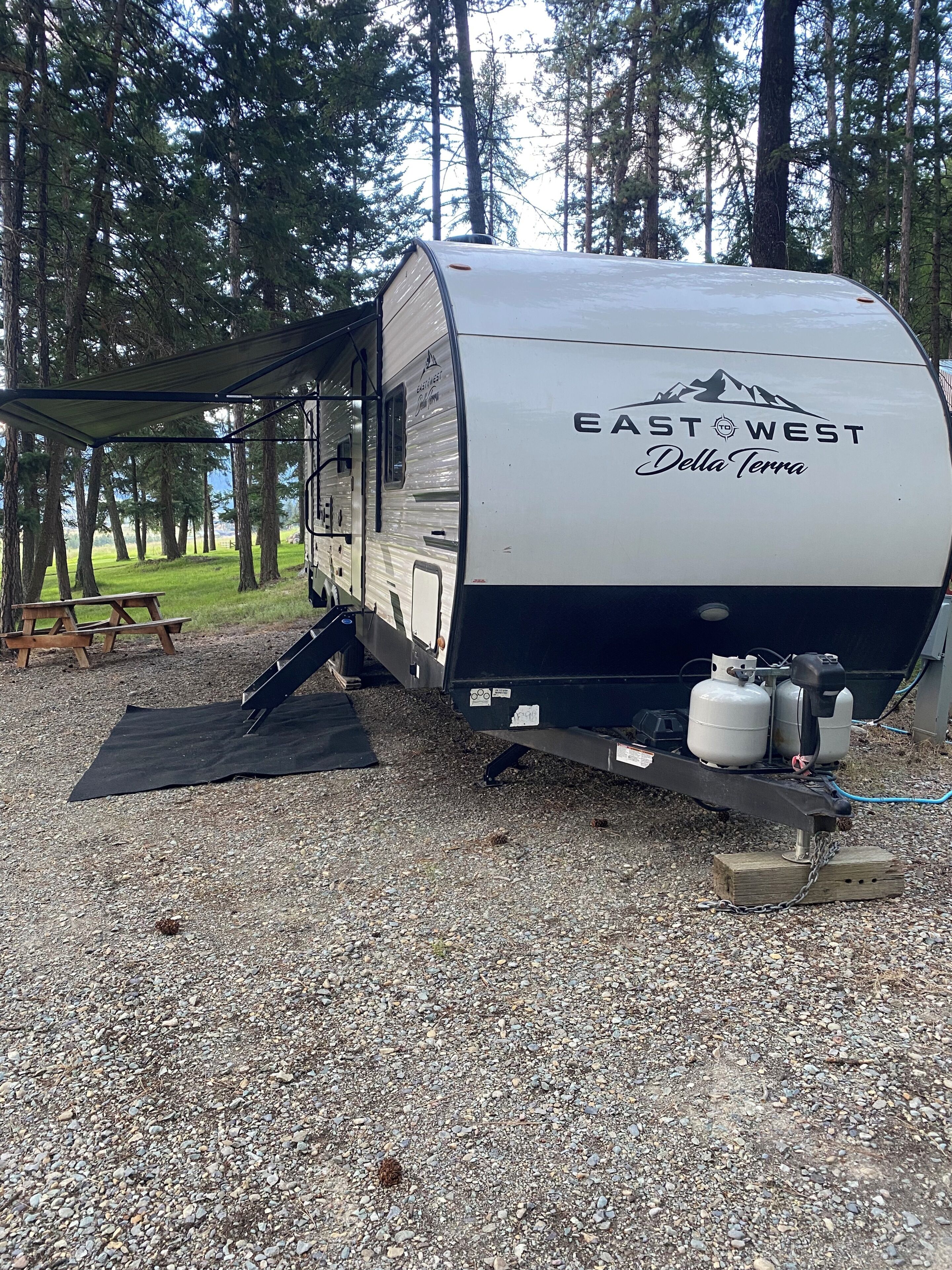 A recreational vehicle with 'EAST WEST Della Terra' branding, a picnic table, and propane tanks.