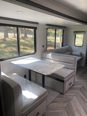 A compact RV interior with a dining table, seating area, and windows offering a view of the outdoors. - Cozy  1-bedroom recreational vehicle with views close to Kalispell. (Kalispell)