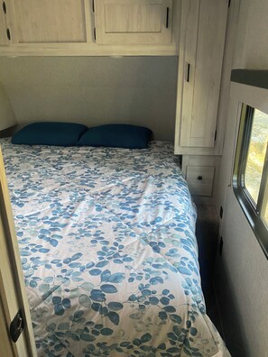 A bed with a floral bedspread, a window on the right, and a door on the left. - Cozy  1-bedroom recreational vehicle with views close to Kalispell. (Kalispell)
